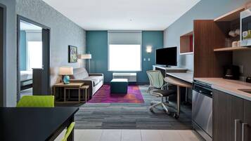 A modern hotel room with a sofa, a dining table, a kitchen area, and a large window providing natural light.