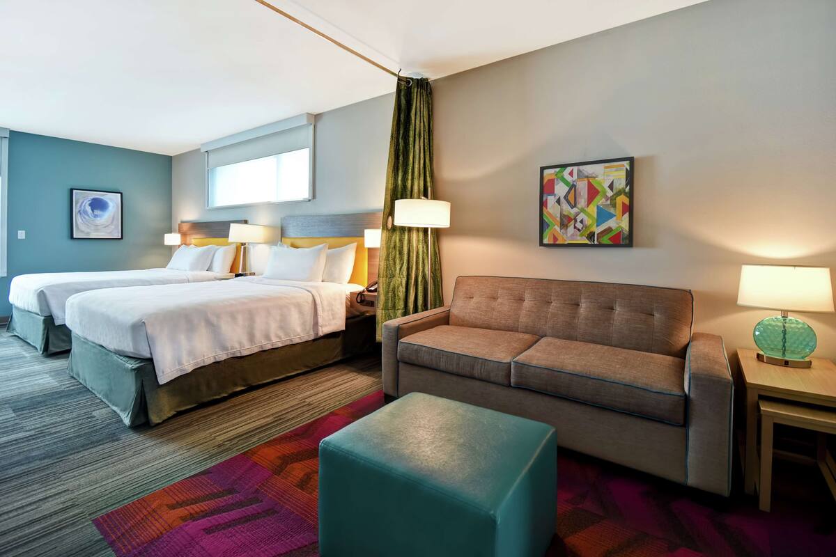 Une chambre d’hôtel avec deux lits, un canapé, une petite table et un tableau abstrait aux couleurs vives accroché au mur.