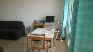 A living room with a black sofa, a wooden dining table with chairs, a television, and a window with curtains. - ANTIBES - LARGE STUDIO BRIGHT, SEA VIEW for 4 pers (Antibes)