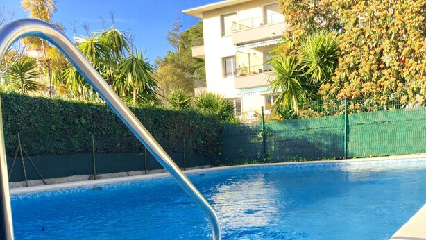 A swimming pool with a metal ladder, surrounded by greenery and a building in the background. - ANTIBES - LARGE STUDIO BRIGHT, SEA VIEW for 4 pers (Antibes)