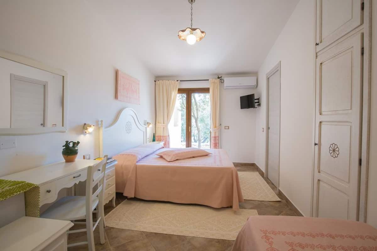 A bedroom with a bed, a desk, a chair, a dresser, a window with curtains, and a wall-mounted TV.