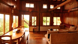 A wooden interior with a dining table, chairs, a sofa, and large windows offering a view of the outdoors. - Mount Totumas Cloud Forest (Volcan)