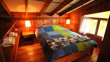 Un dormitorio de cabaña de madera con una cama, dos lámparas de noche, un banco con toallas y una ventana con cortinas.
