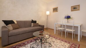 Una sala de estar con sofá, una mesa de centro de vidrio, una mesa de comedor con sillas, una lámpara de pie y un tapete estampado.