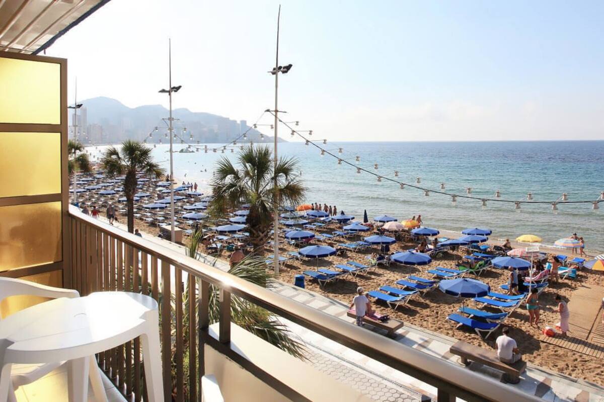A beach with rows of blue umbrellas and sun loungers, a wooden deck, and a view of the sea and distant mountains.