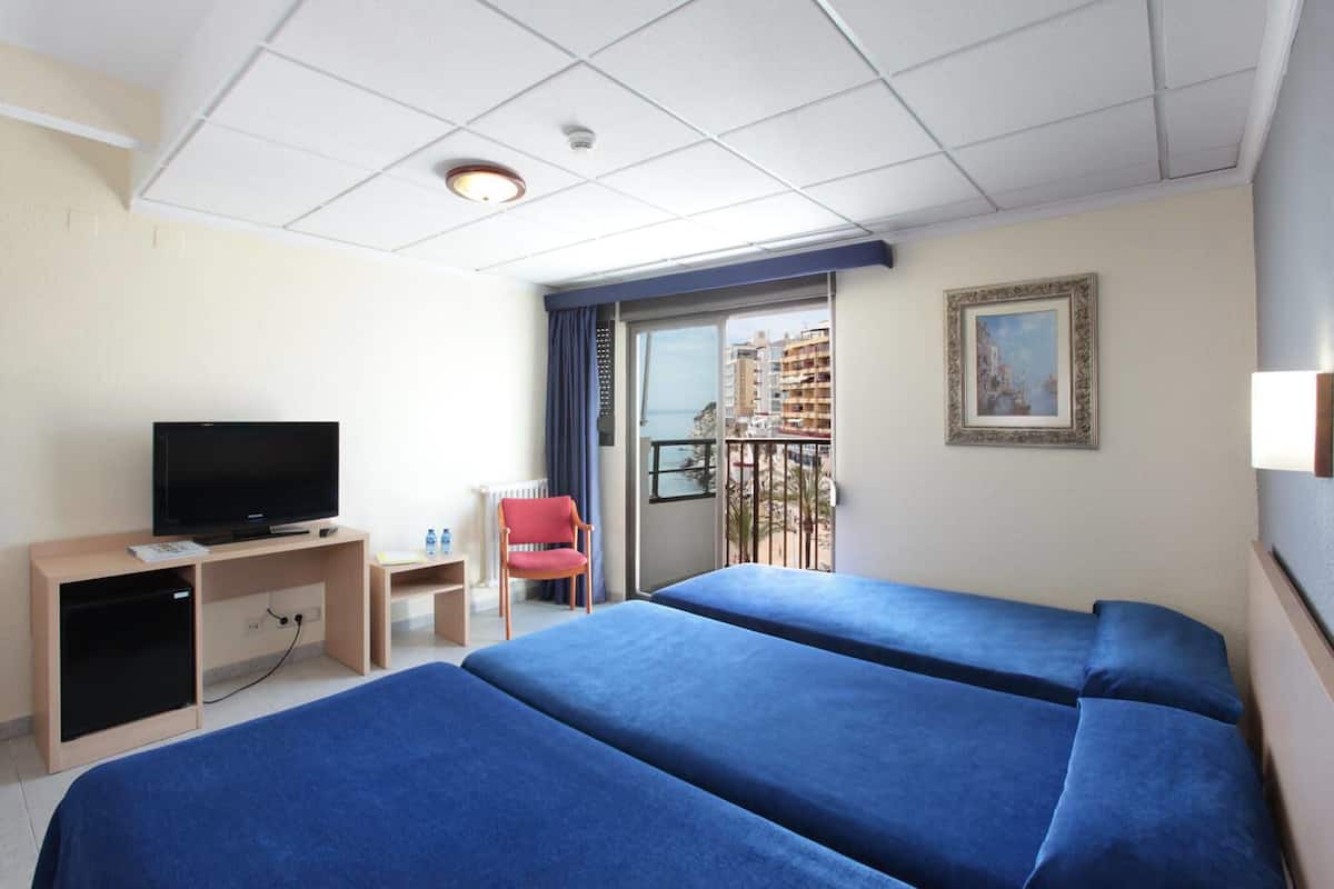 A hotel room with two beds, a TV, a desk, a chair, and a balcony with a view of buildings.