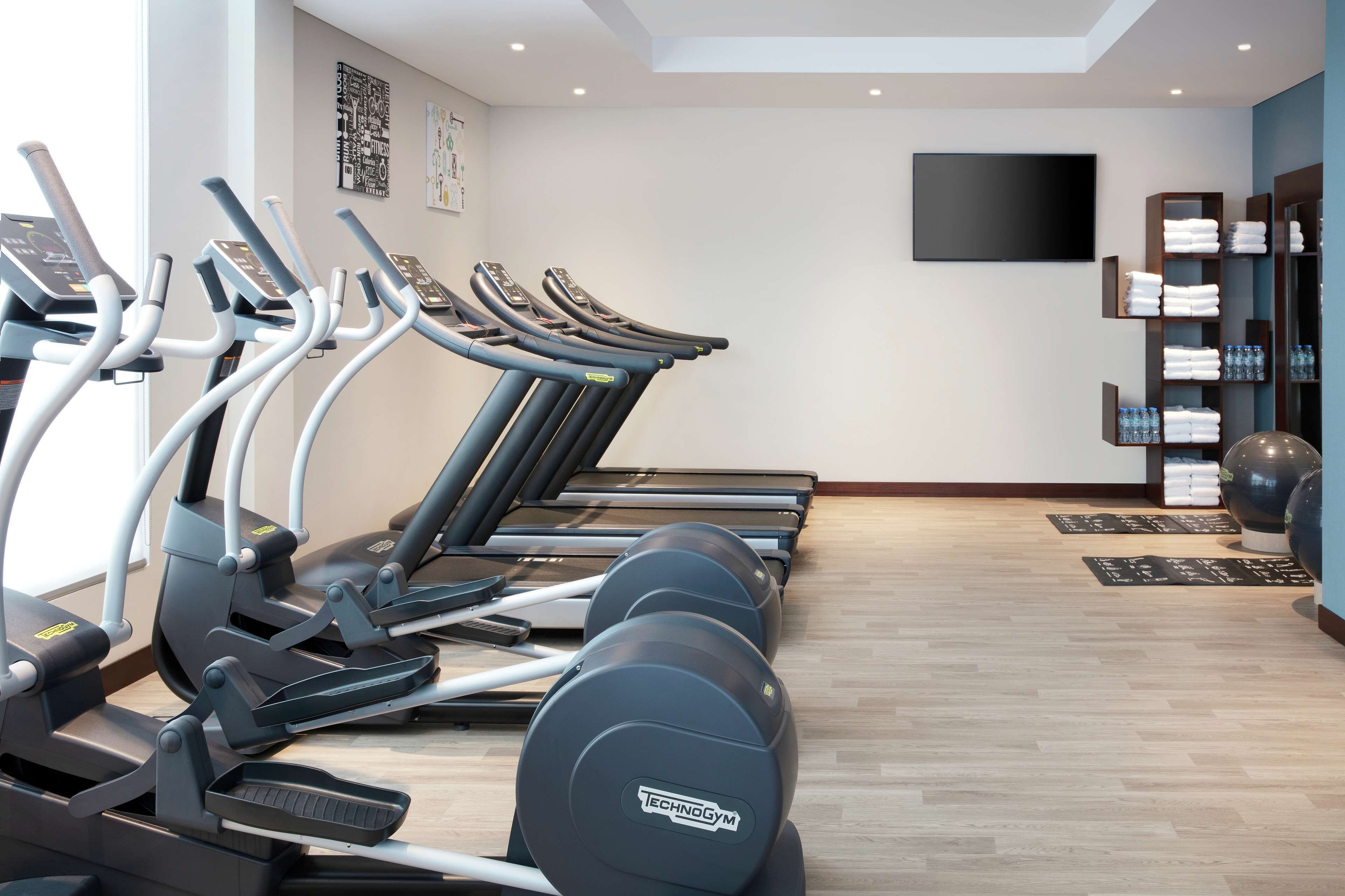 Un gimnasio con varias cintas de correr, un televisor de pantalla plana y toallas y botellas de agua ordenadas.