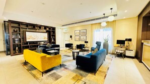 A modern living room with a large flat-screen TV, a yellow sofa, a blue sofa, and a coffee table. There are multiple chairs and a small table with a lamp. The room has a large window with curtains and a plant.
