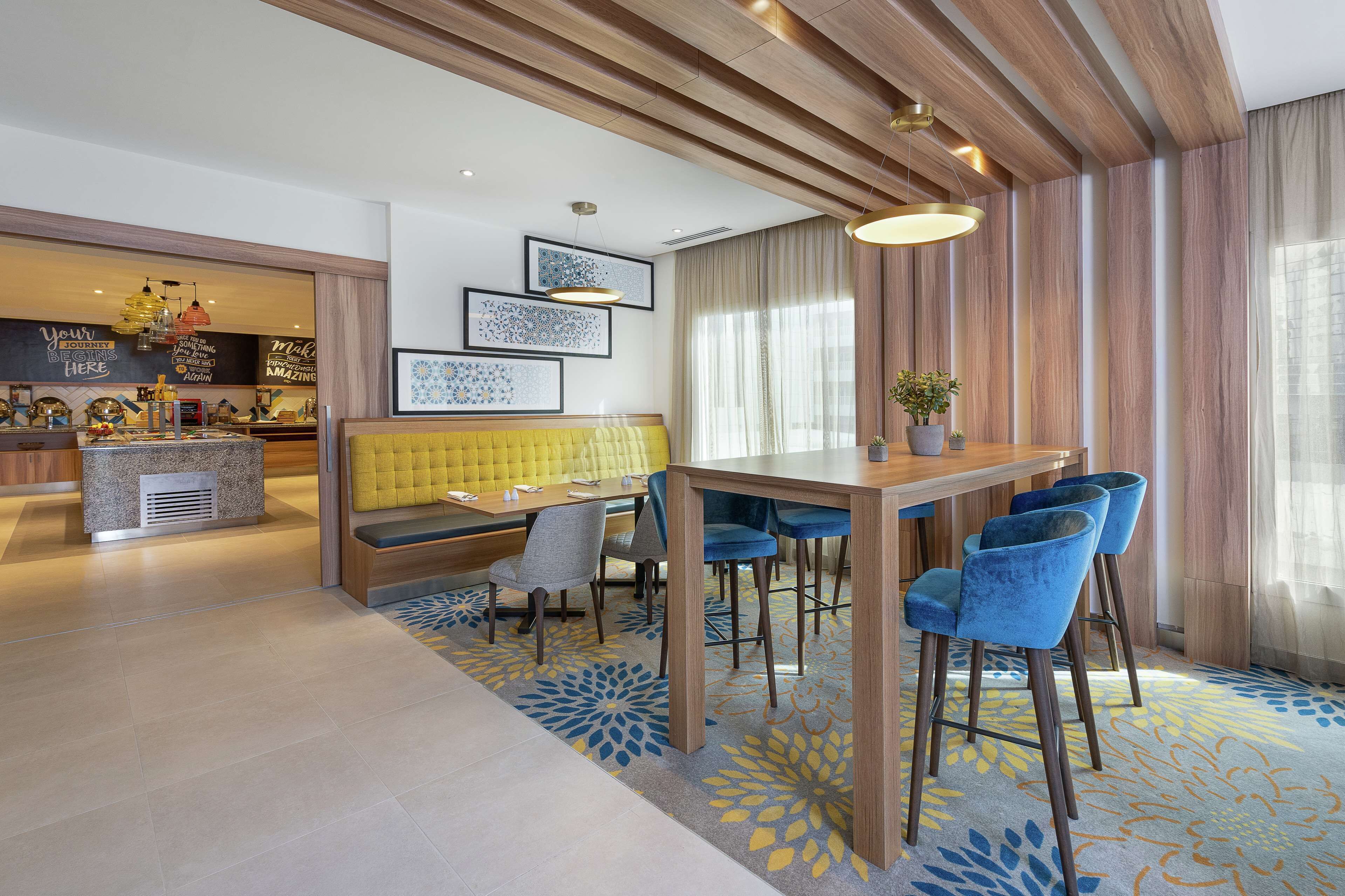 Een moderne hotel lobby met een houten plafond, een gele bank, een houten eettafel met blauwe stoelen en lijstwerken aan de muren.