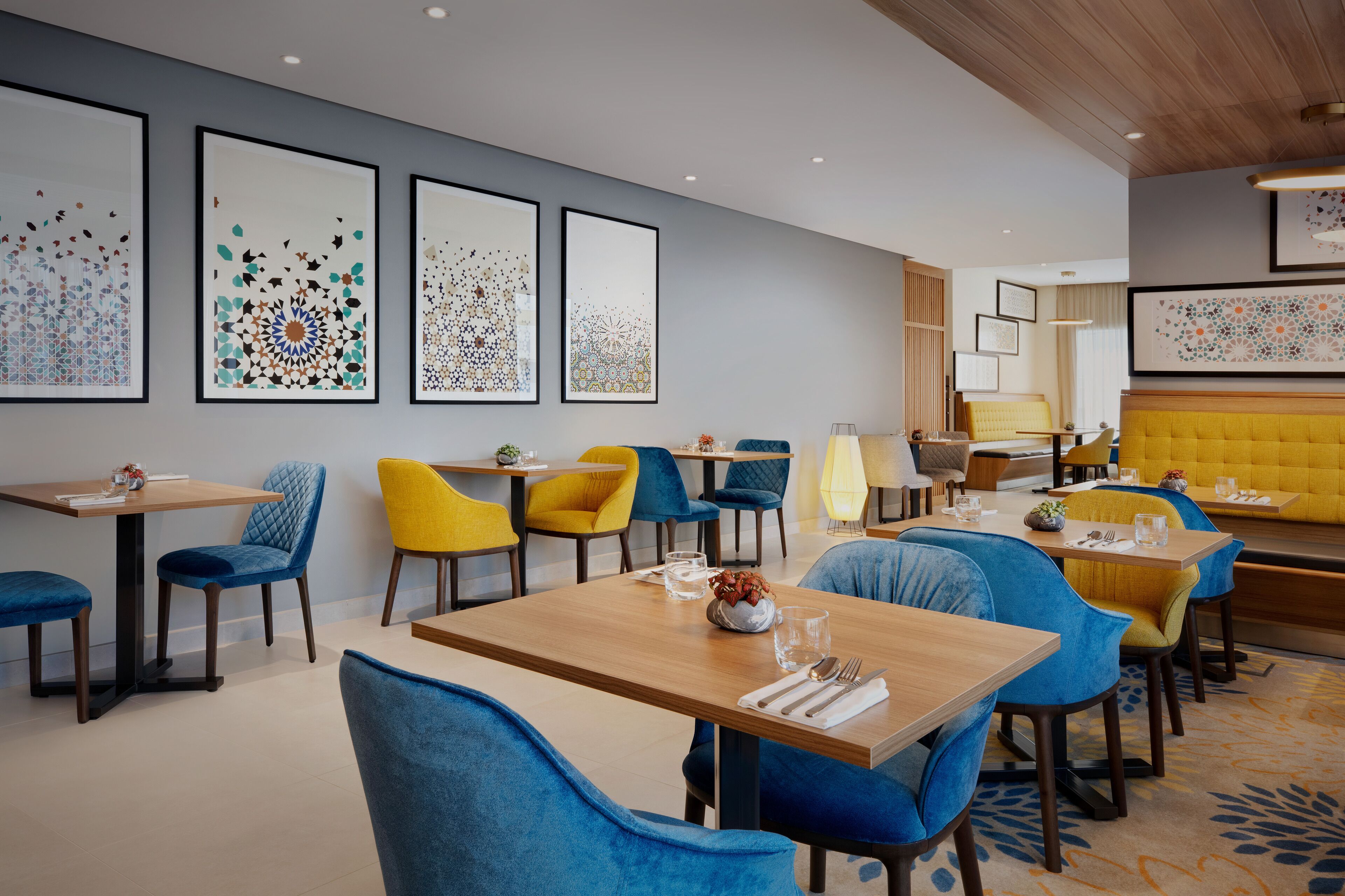 A modern dining area with multiple tables and chairs, framed artwork on the walls, and a yellow cushioned bench in the background.