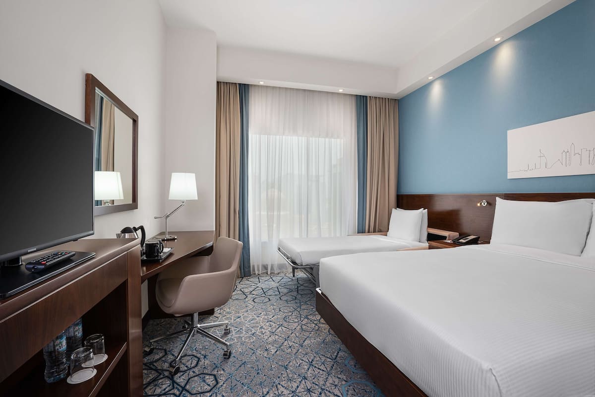 Habitación de hotel con una cama grande, un escritorio con televisor, una silla y una ventana con cortinas.