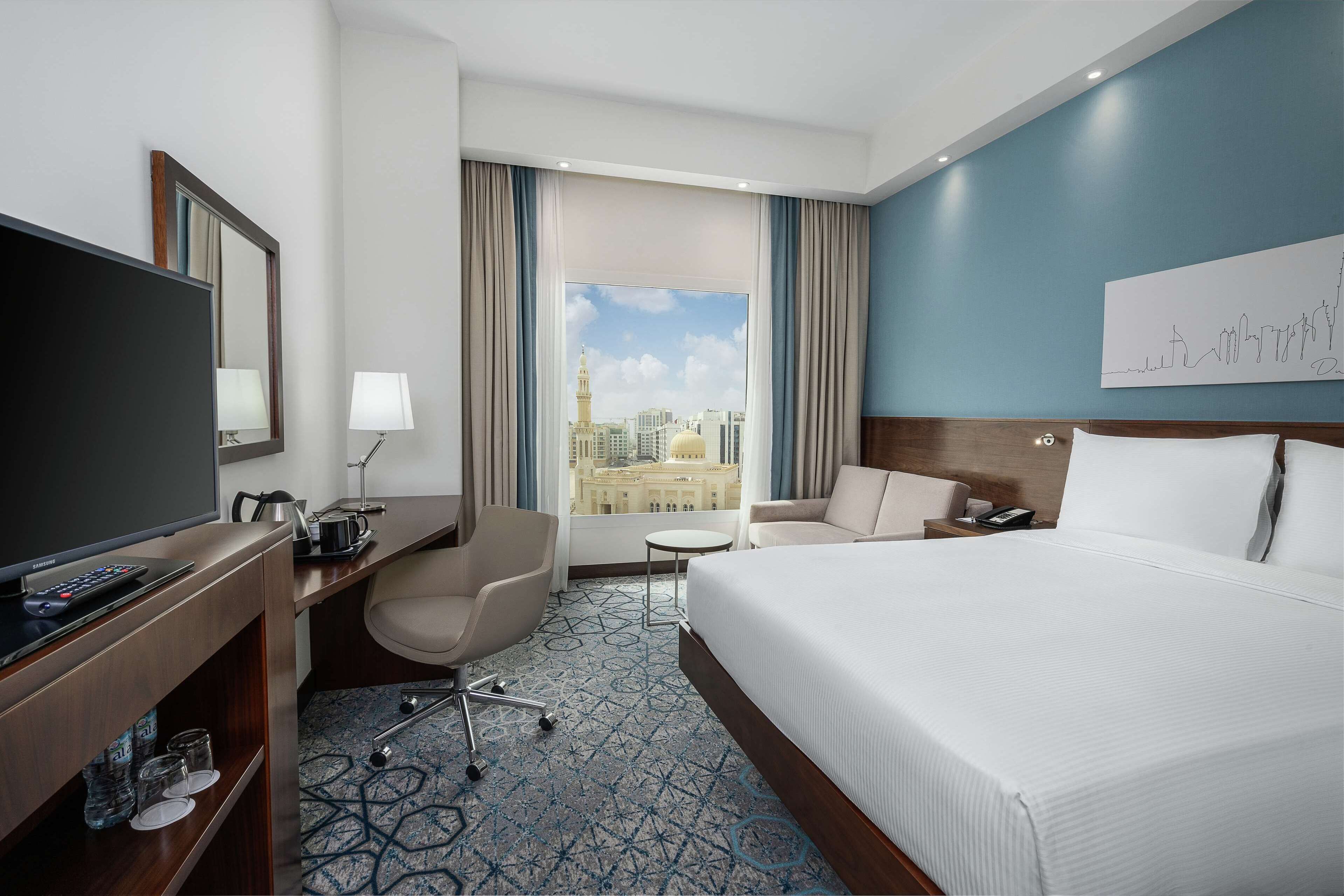 A hotel room with a large bed, a desk with a television, a chair, a small table, and a view of a cityscape through the window.