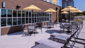 An outdoor patio with tables and chairs, large windows, and yellow umbrellas. - Hilton Garden Inn Kansas City Airport (Kansas City)