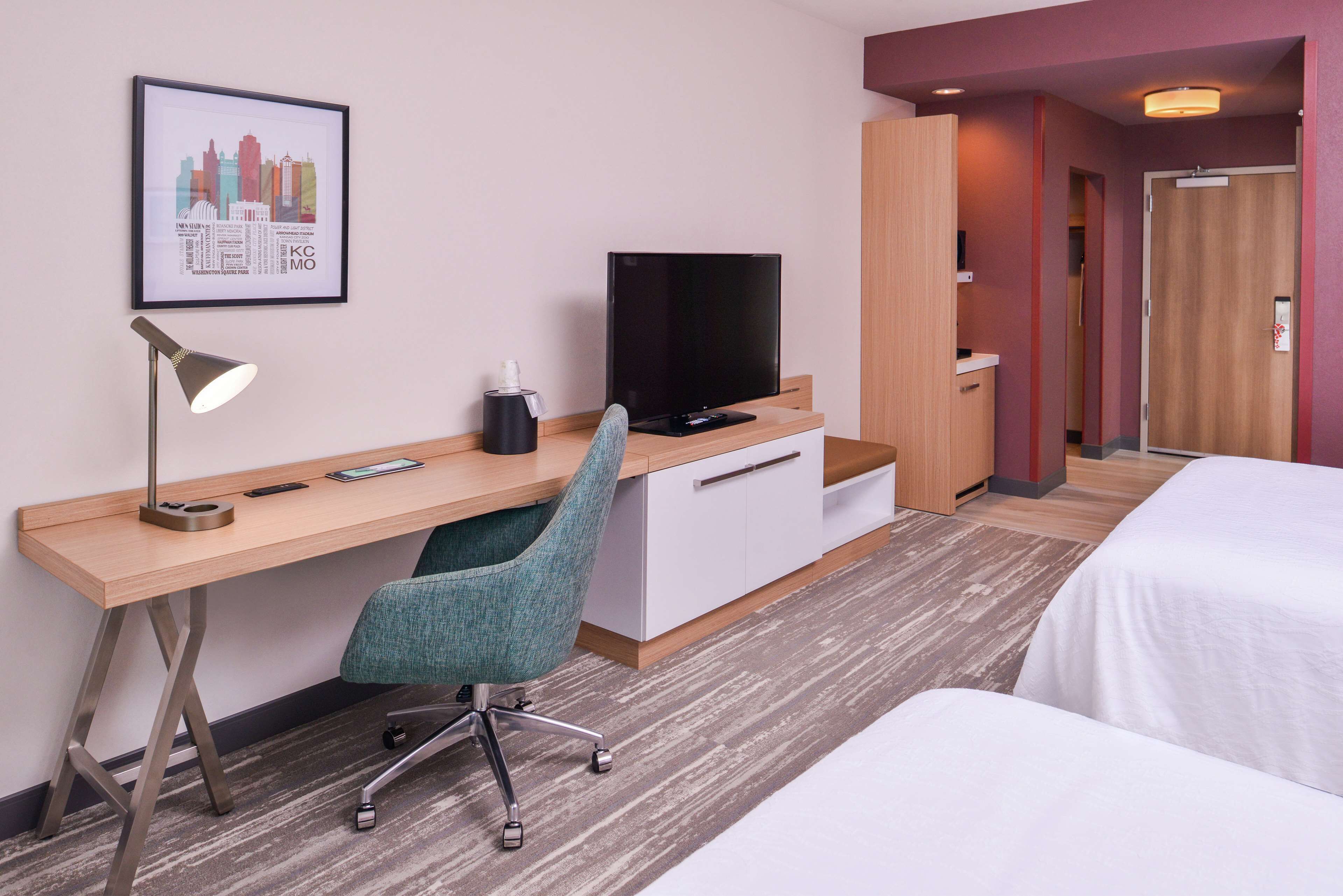 Habitación de hotel con escritorio de madera, una silla de oficina color verde azulado, televisor de pantalla plana, mesita de noche y cama.