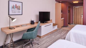 Habitación de hotel con escritorio de madera, una silla de oficina color verde azulado, televisor de pantalla plana, mesita de noche y cama.