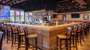 A bar area with a long counter, multiple taps, and a selection of bottles. There are several wooden bar stools arranged around the counter. - Hilton Garden Inn Kansas City Airport (Kansas City)