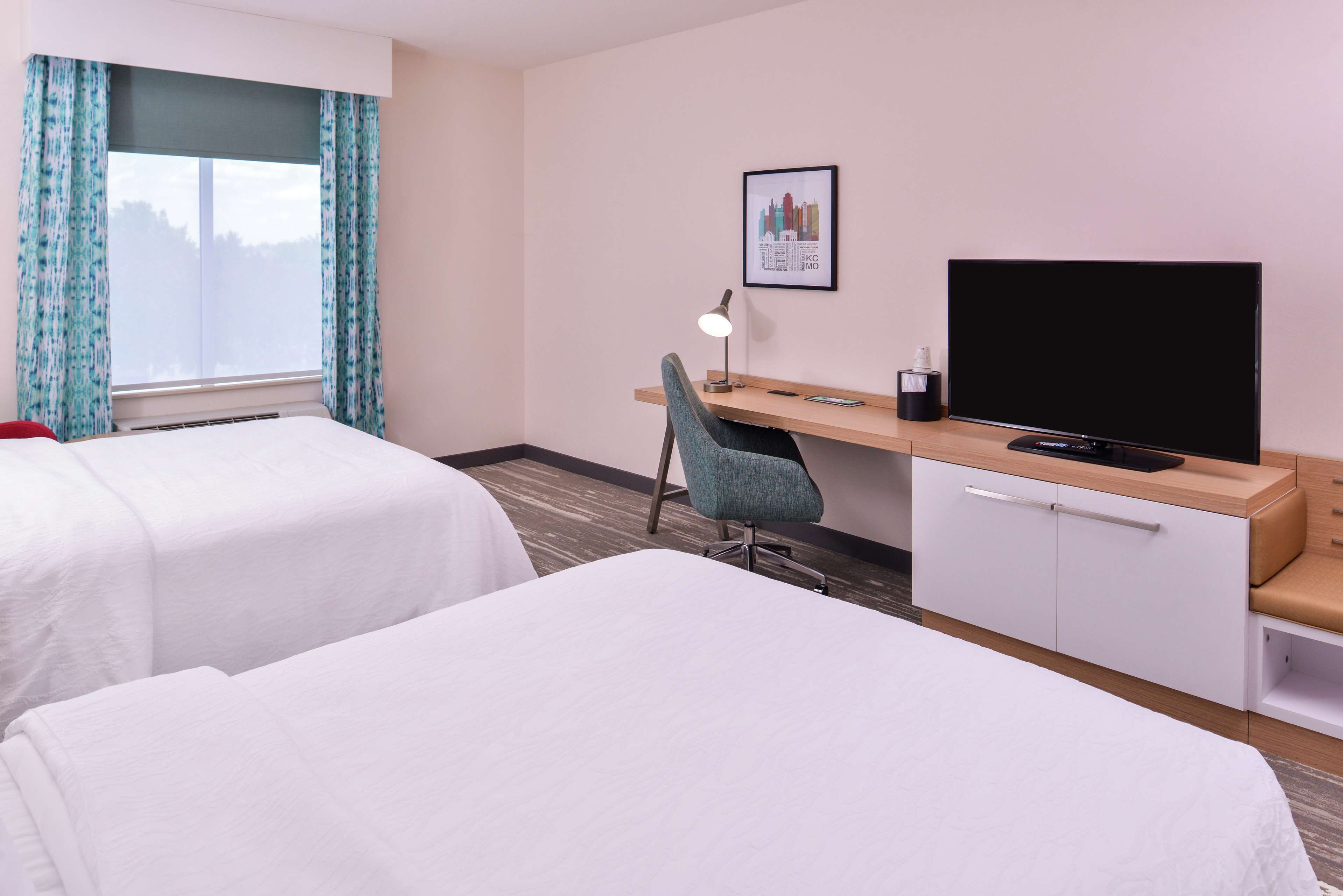 Hotellihuone, jossa on kaksi sänkyä, työpöytä tuolilla, televisio ja seinällä oleva kehystetty kuva.