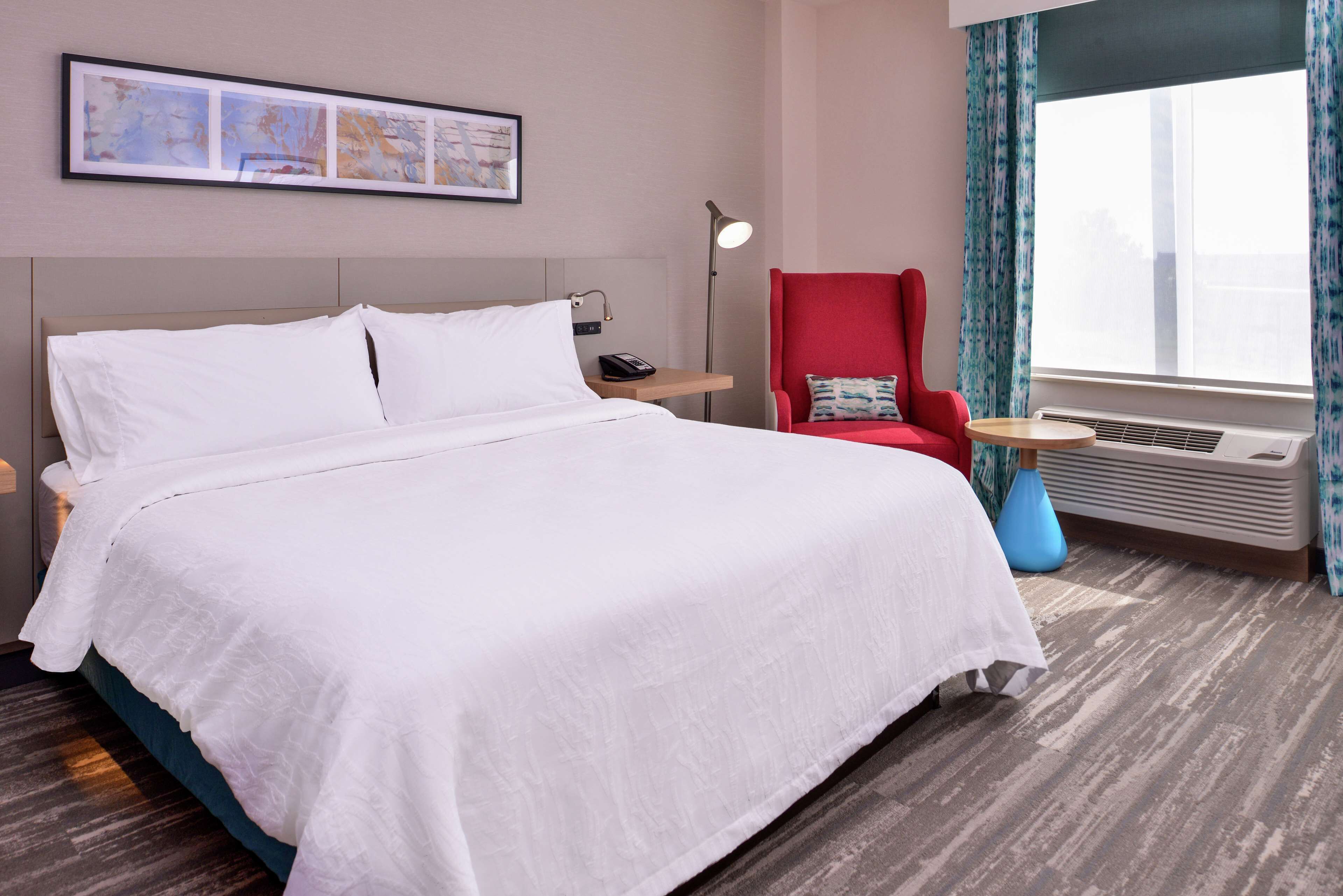Ein Hotelzimmer mit einem großen Bett, einem Schreibtisch mit Lampe, einem roten Sessel und einem großen Fenster mit Vorhängen.
