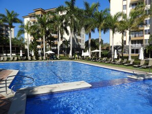 A swimming pool with lounge chairs and umbrellas, surrounded by palm trees and a multi-story building.