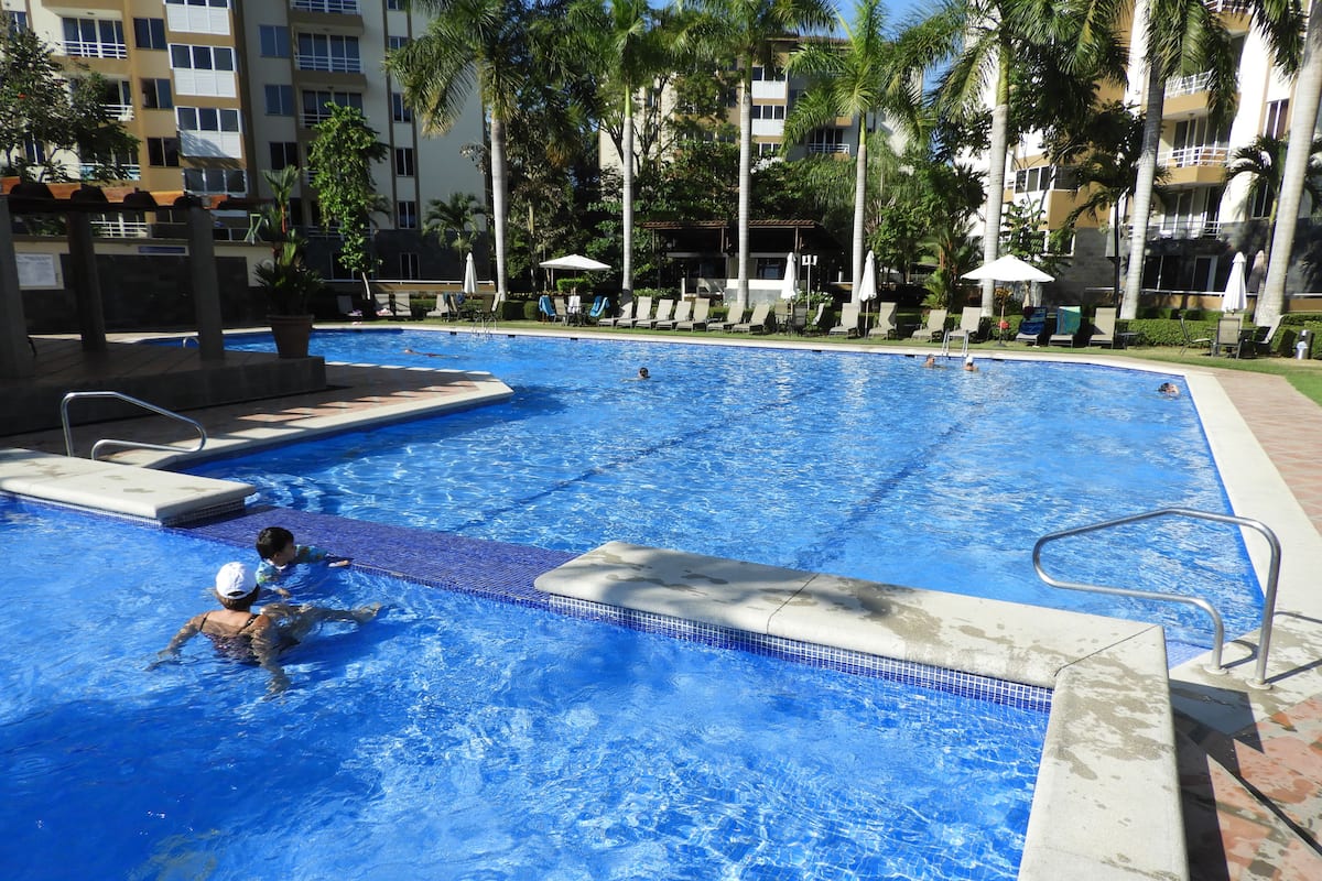A swimming pool with lounge chairs and umbrellas, surrounded by palm trees and a multi-story building.