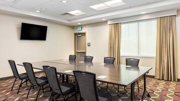A conference room with a long table, six chairs, a flat-screen TV, an exit sign, and a window with curtains.