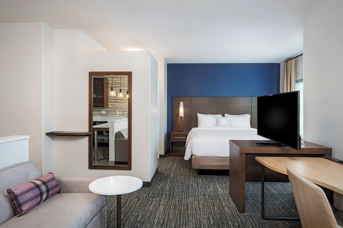 A hotel room with a bed, a TV, a desk, a chair, a small table, a sofa, and a mirror.