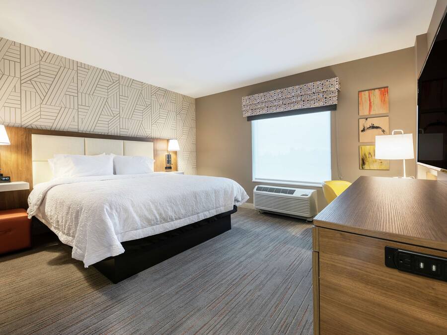 Ein modernes Hotelzimmer mit einem großen Bett, Nachttischlampen, einer fest installierten Heizung, einem großen Fenster und einem Flachbildfernseher.