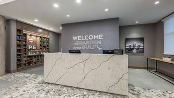 Un mostrador de recepción con encimera de mármol y un cartel que dice “Welcome to the Garden of the Gulf”. Hay dos portátiles HP sobre el mostrador, y una estantería grande con varios objetos, incluyendo un cartel con la inscripción “PE”. El suelo tiene un diseño de mosaico.