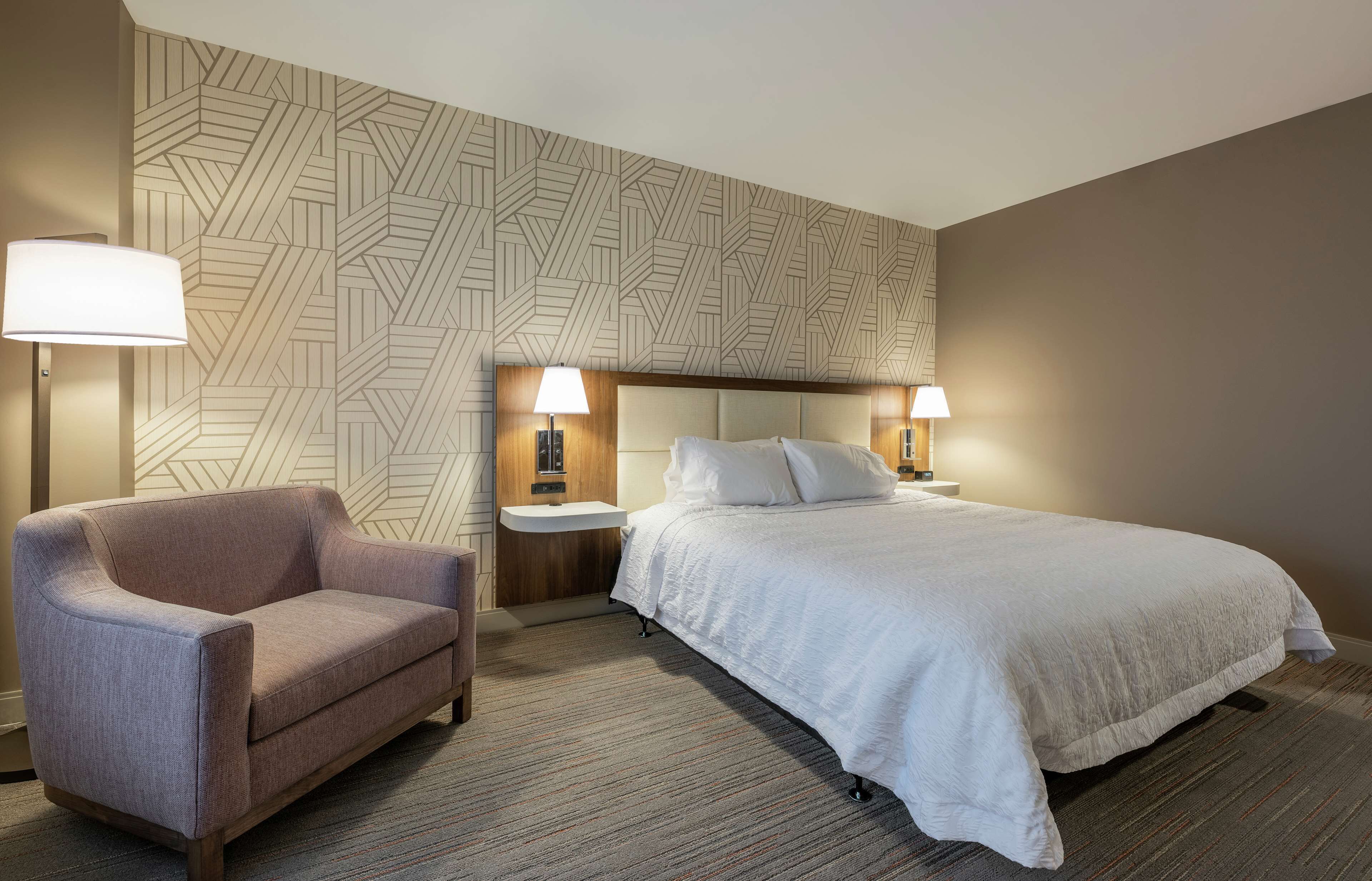 A hotel room with a bed, a chair, two bedside tables with lamps, and a patterned wall.