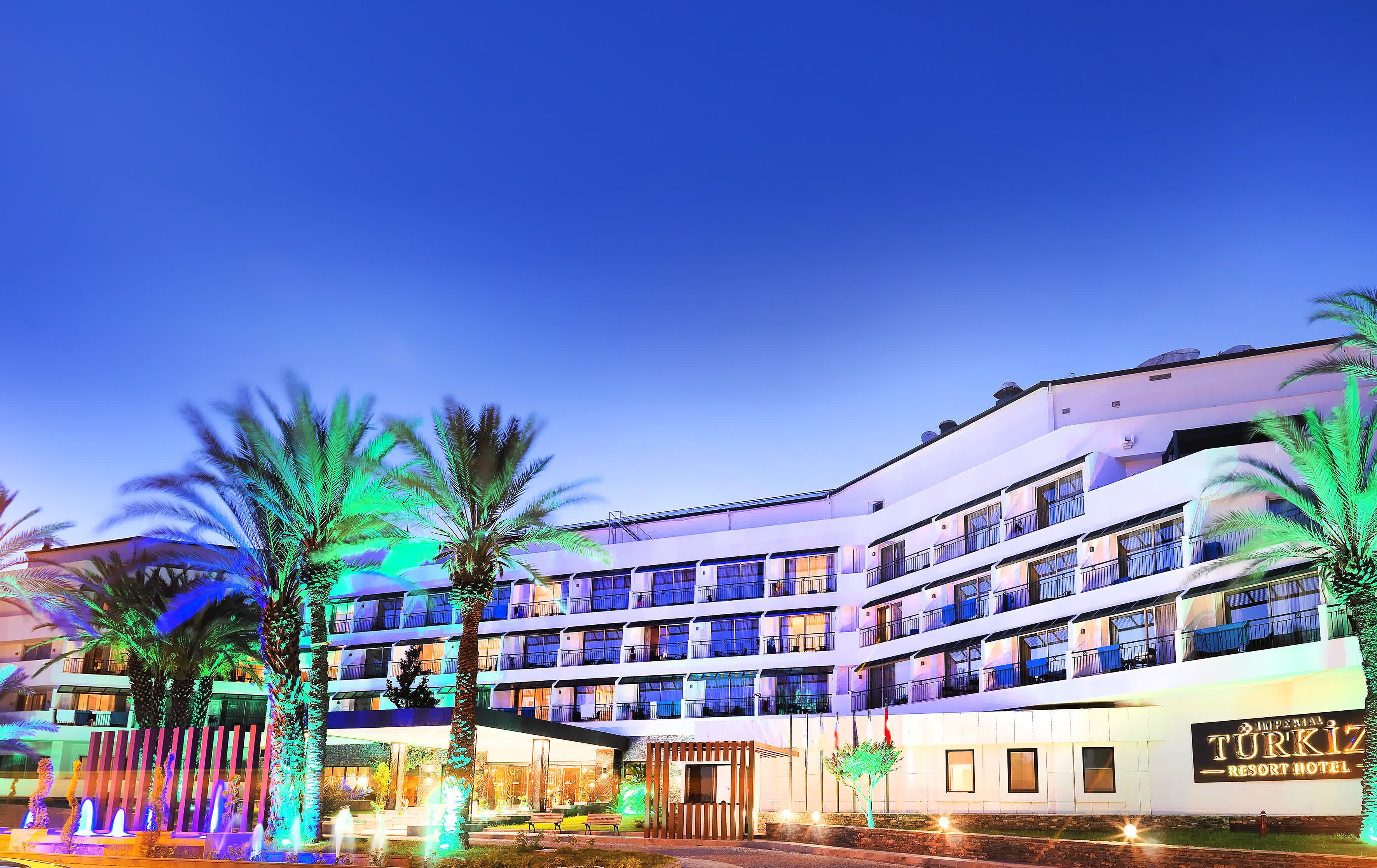 Et hotel med flere etager og altaner, et skilt med "Imperial Turkish Resort Hotel" og et veloplyst indgangsparti med palmer.
