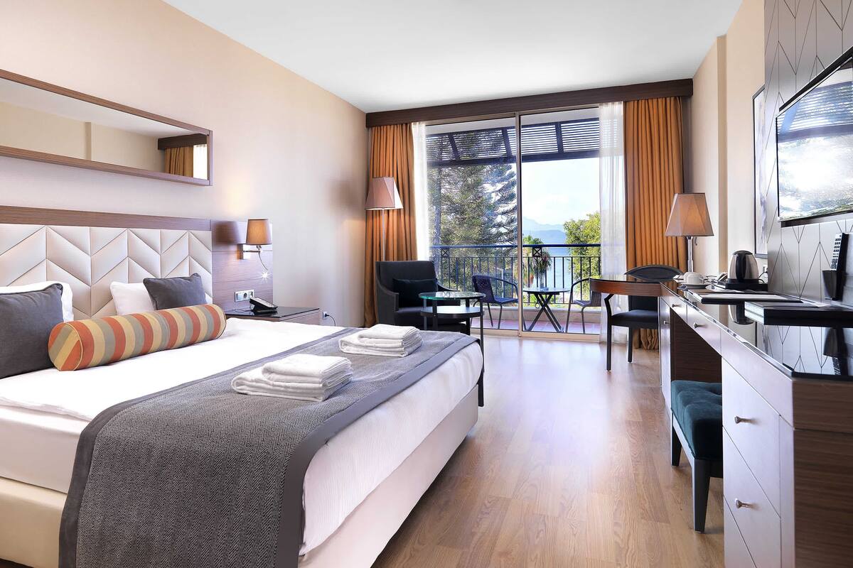 A hotel room with a large bed, a desk, a chair, a small table, and a balcony with a view.