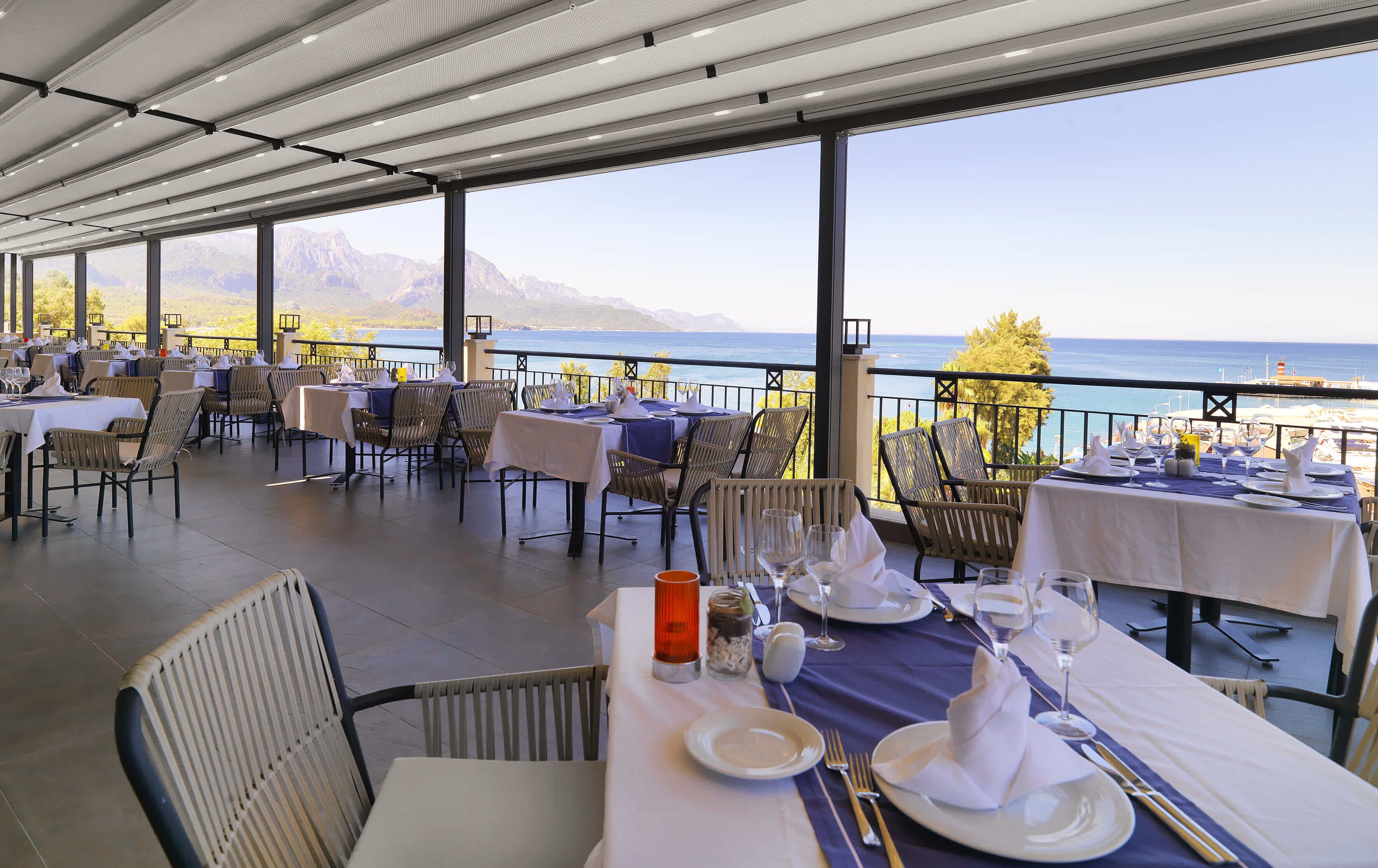 A restaurant with tables set for dining, overlooking a scenic view of the sea and mountains.