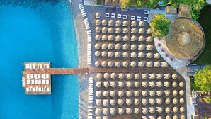An aerial view of a beachside resort with multiple rows of sun loungers and umbrellas, a wooden pier, and a circular seating area with a central fountain.