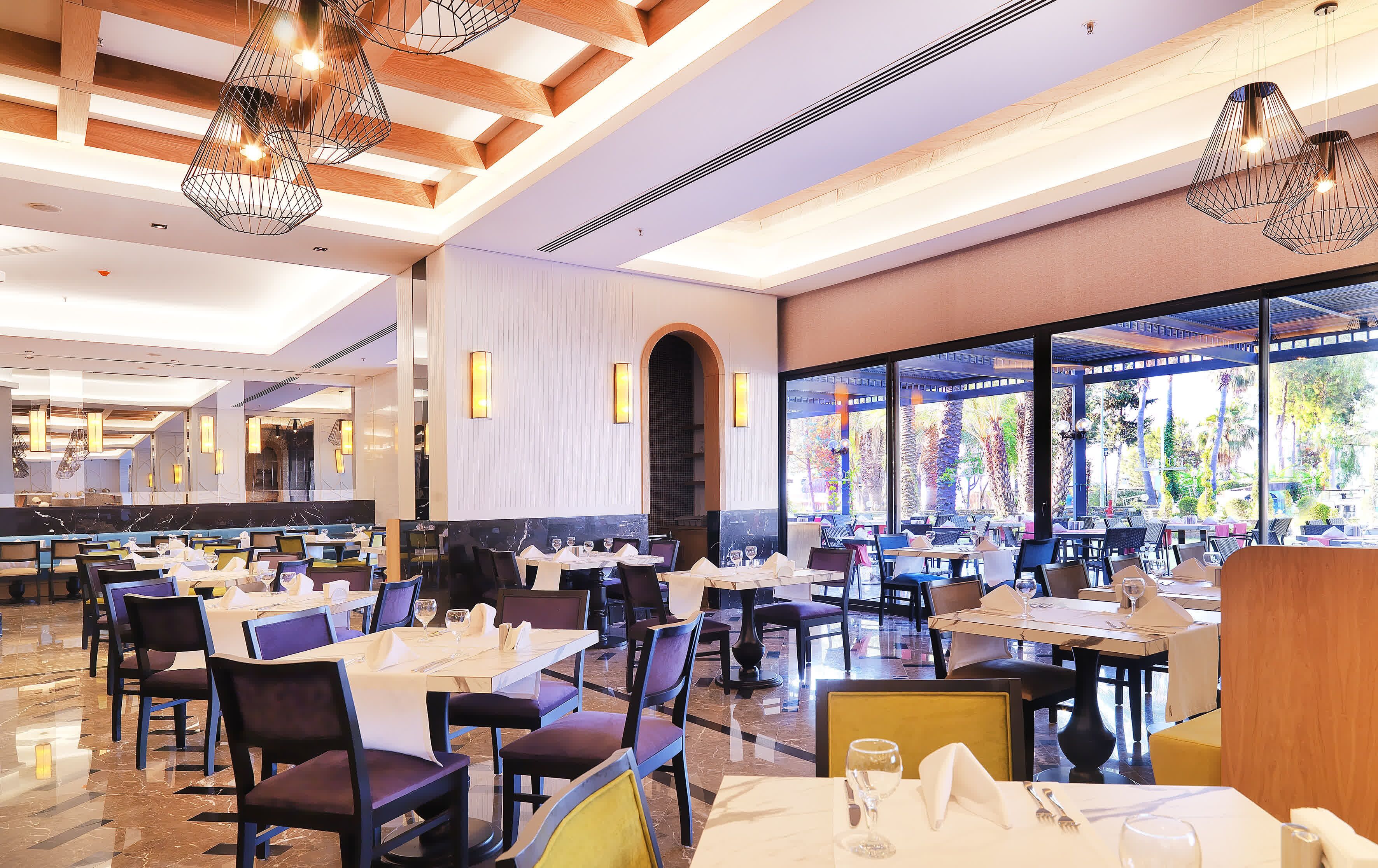 A spacious restaurant with multiple dining areas, modern lighting fixtures, and large windows offering views of the outdoors.