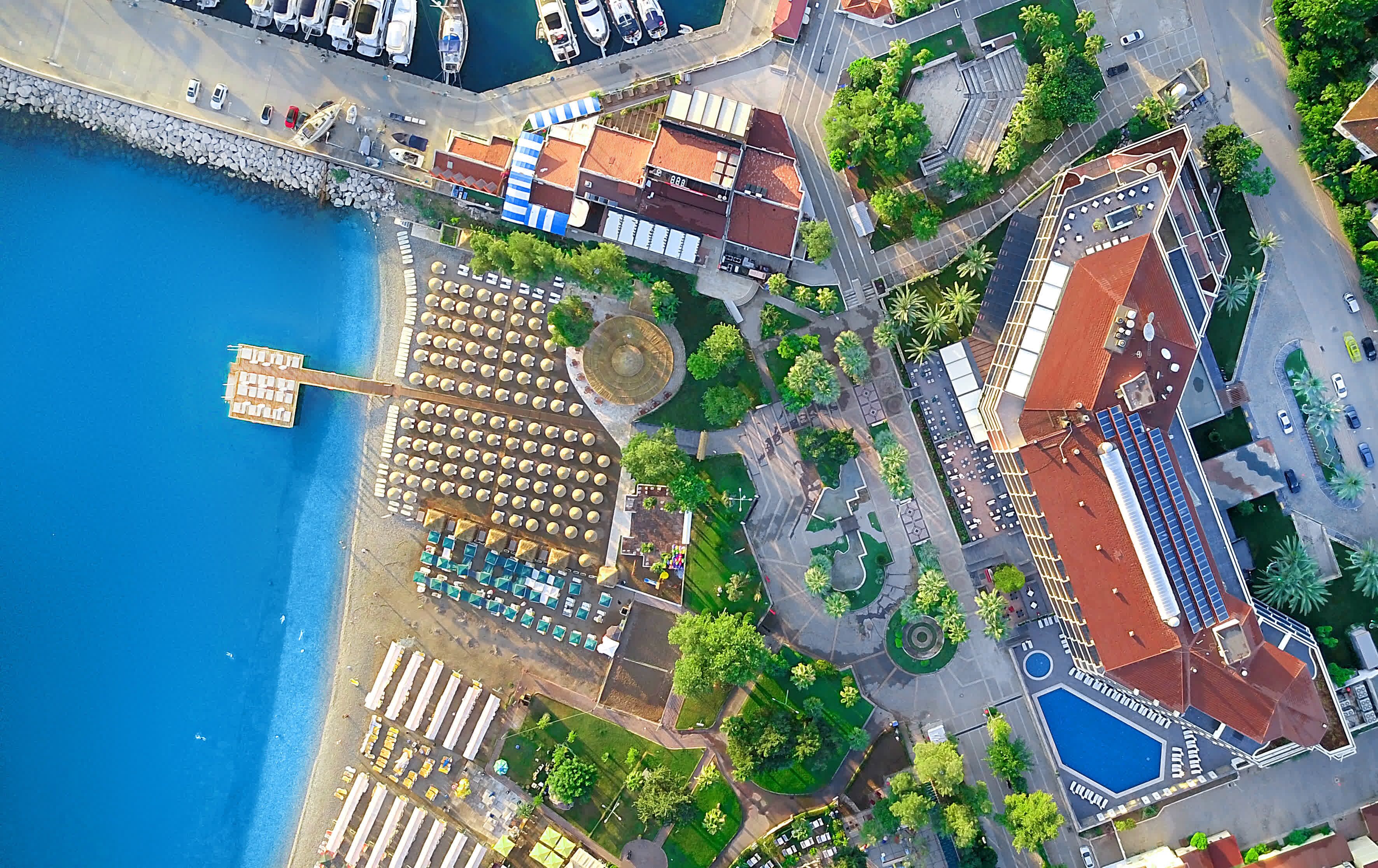 An aerial view of a coastal area with a marina, beach with sun loungers, and a building with a red roof.