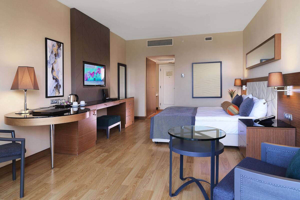A hotel room with a bed, a desk, a chair, a small table, and a TV.