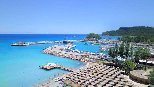 Un port de plaisance situé sur la côte, avec des bateaux, une jetée et une zone de plage équipée de parasols et de chaises longues.