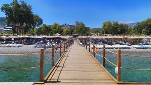 Une jetée en bois qui mène à une plage où s’alignent des rangées de transats et de parasols.