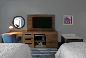 A hotel room with a wooden TV stand, a flat-screen TV, a desk with a lamp, a microwave, and a small bedside table. - Hampton Inn Weslaco (Weslaco)