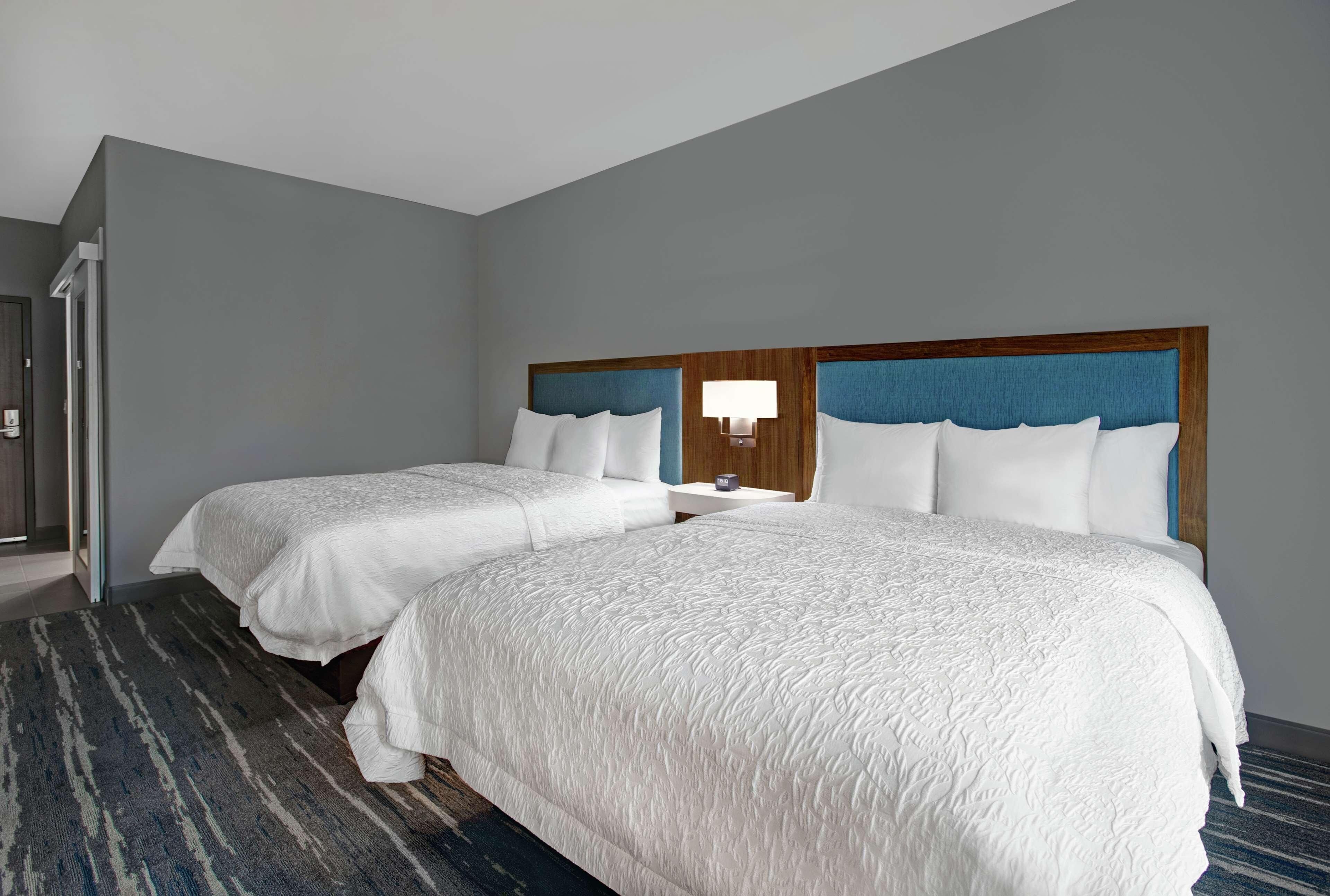 A hotel room with two beds, a blue headboard, a bedside table with a lamp, and a striped carpet.