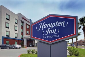En skylt för Hampton Inn by Hilton framför en byggnad med många fönster och en röd och blå fasad.