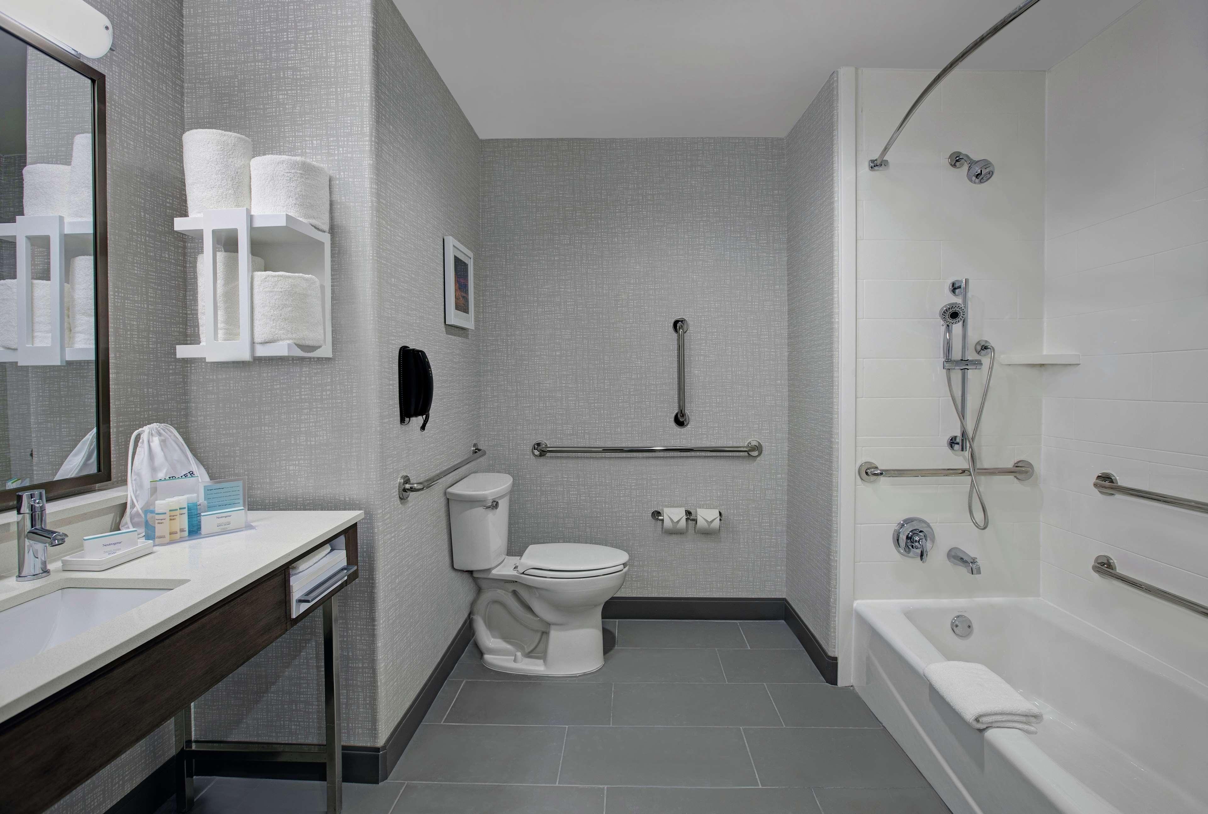 A modern bathroom with a toilet, sink, and bathtub. It includes a handrail, grab bars, and a showerhead.