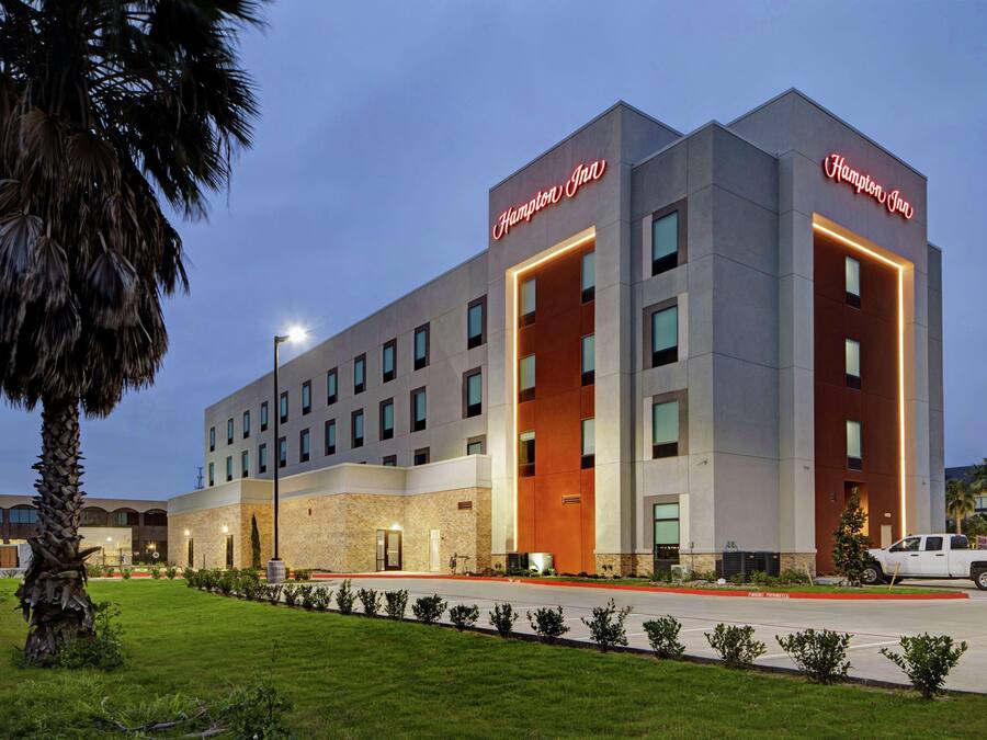 A modern Hampton Inn hotel with a prominent sign, a well-maintained lawn, and a parking area.