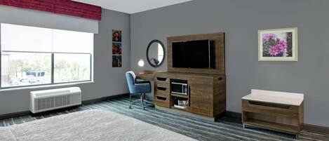 A modern bedroom with a large bed, a desk, a TV, and a window with a view of trees.