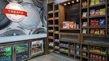Zona de exhibición de bocadillos con papas fritas y aperitivos varios, una sección refrigerada con bebidas, y una pizarra con un mensaje de bienvenida.