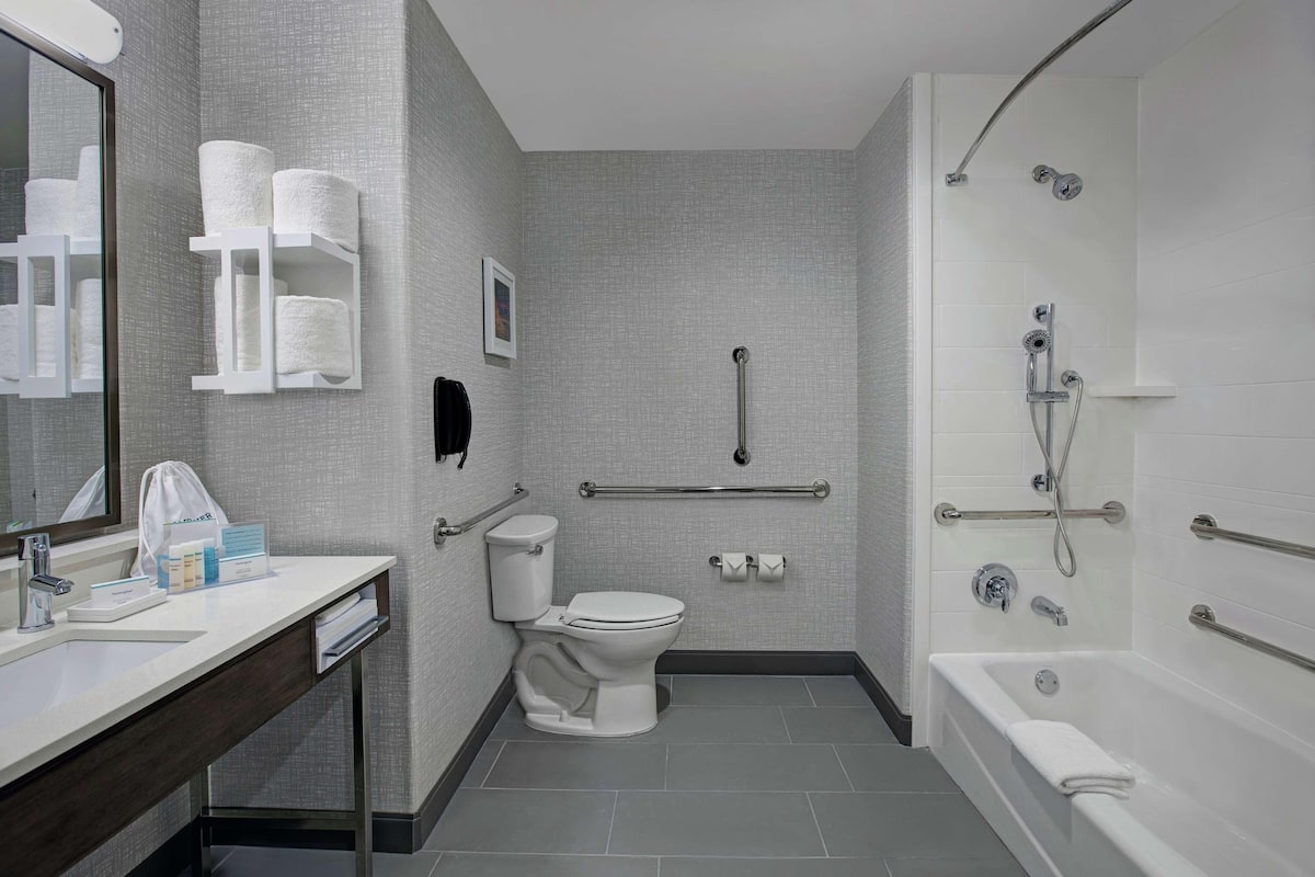 Une salle de bain moderne équipée de toilettes, d’un lavabo et d’une baignoire. Elle comprend également une barre d’appui, des barres de maintien et un pommeau de douche.