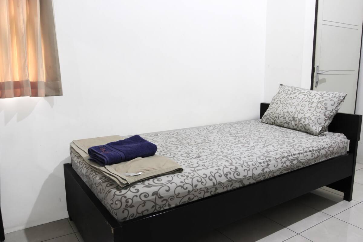 A single bed with a patterned bedspread, a folded towel, and a bag on the bed.