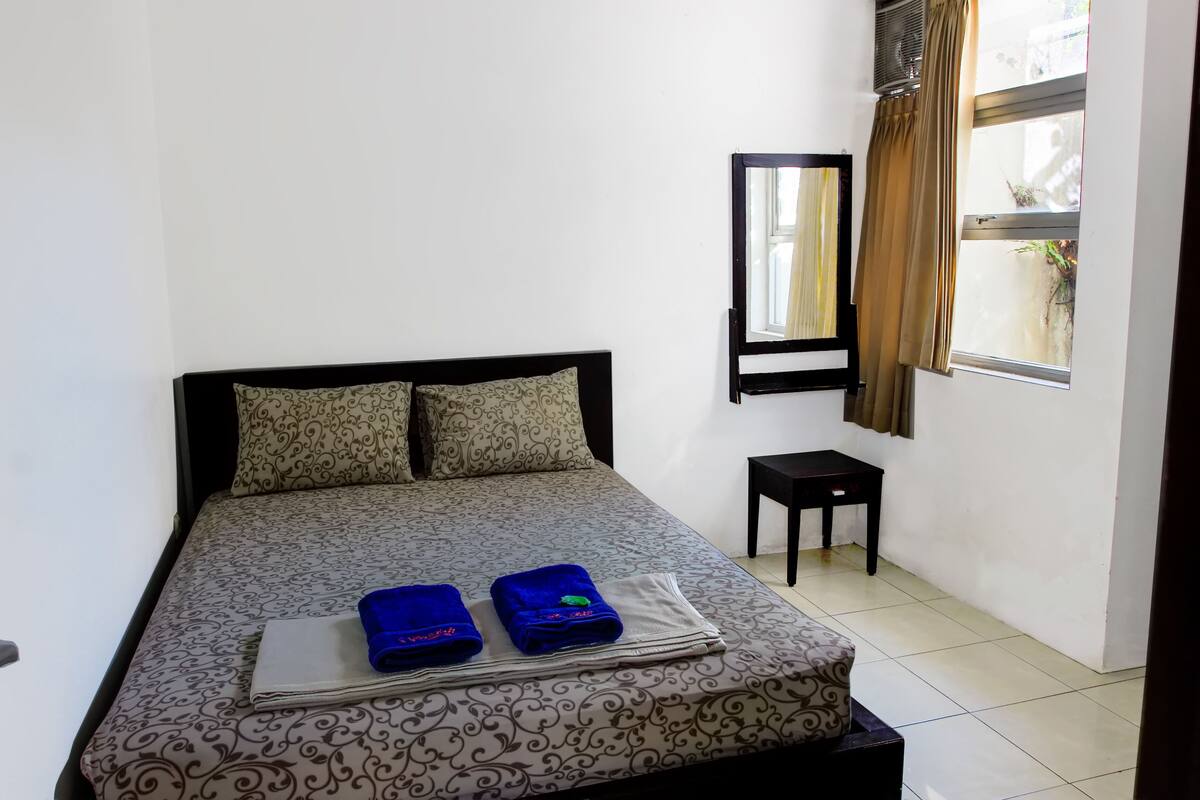 A neatly made bed with patterned bedding and two folded towels. A small table is beside the bed, and a window with curtains is visible.