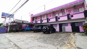 Un immeuble à plusieurs étages, avec une façade violette, un camion bleu garé devant et un VUS noir.