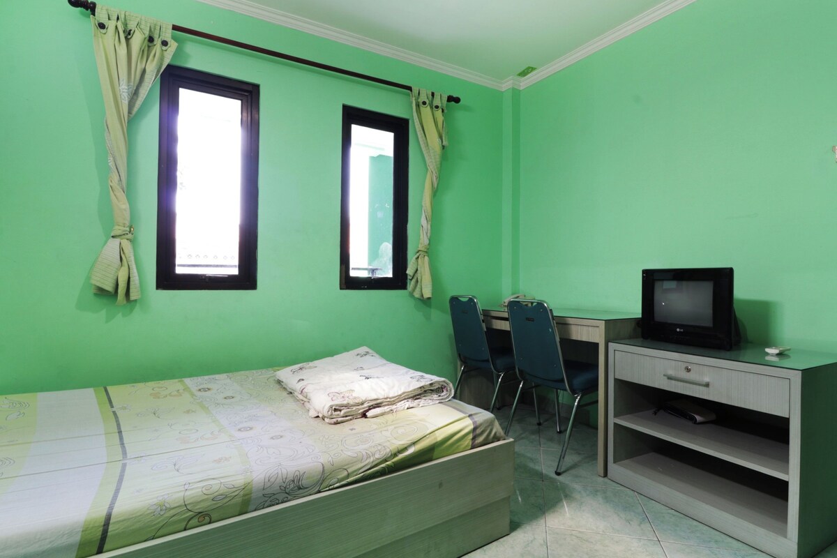 A room with a bed, a desk with a chair, a television, and two windows with curtains.